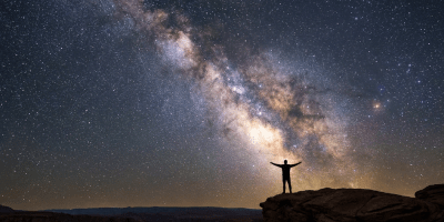 Silhouette of a person with outstretched arms on a cliff under the Milky Way galaxy.